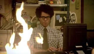 richard ayoade sits at his desk while a fire lights up in the it crowd