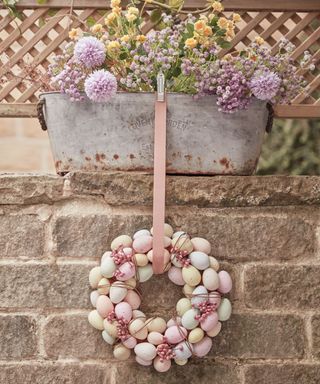 A pastel colored egg wreath hanging on a pink ribbn clipped onto a silver plnter with purple and yellow flowers in it, against a gray brick wall and wooden lattice fence