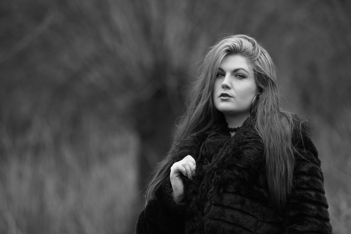 Black and white photograph of a woman, wearing glasses and a black coat, against a blurry outdoor background