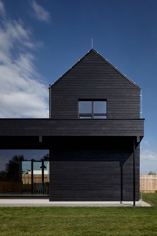 The exterior of a residential home clad in black timber planks with tall pitched-roof unit at one end of the structure surrounded by grey concrete and green lawn