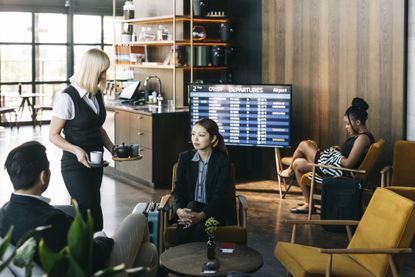 Three travelers lounge in a cafe area of an airport lounge, with a monitor showing flight status. A server brings coffee.