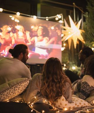 Four people sat outside at nighttime on patterned throw pillows looking to a large outdoor screen with four people projected onto it and a large star light next to it