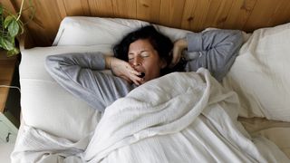 A woman with dark hair lies in bed yawning because she is so tired and ready to sleep