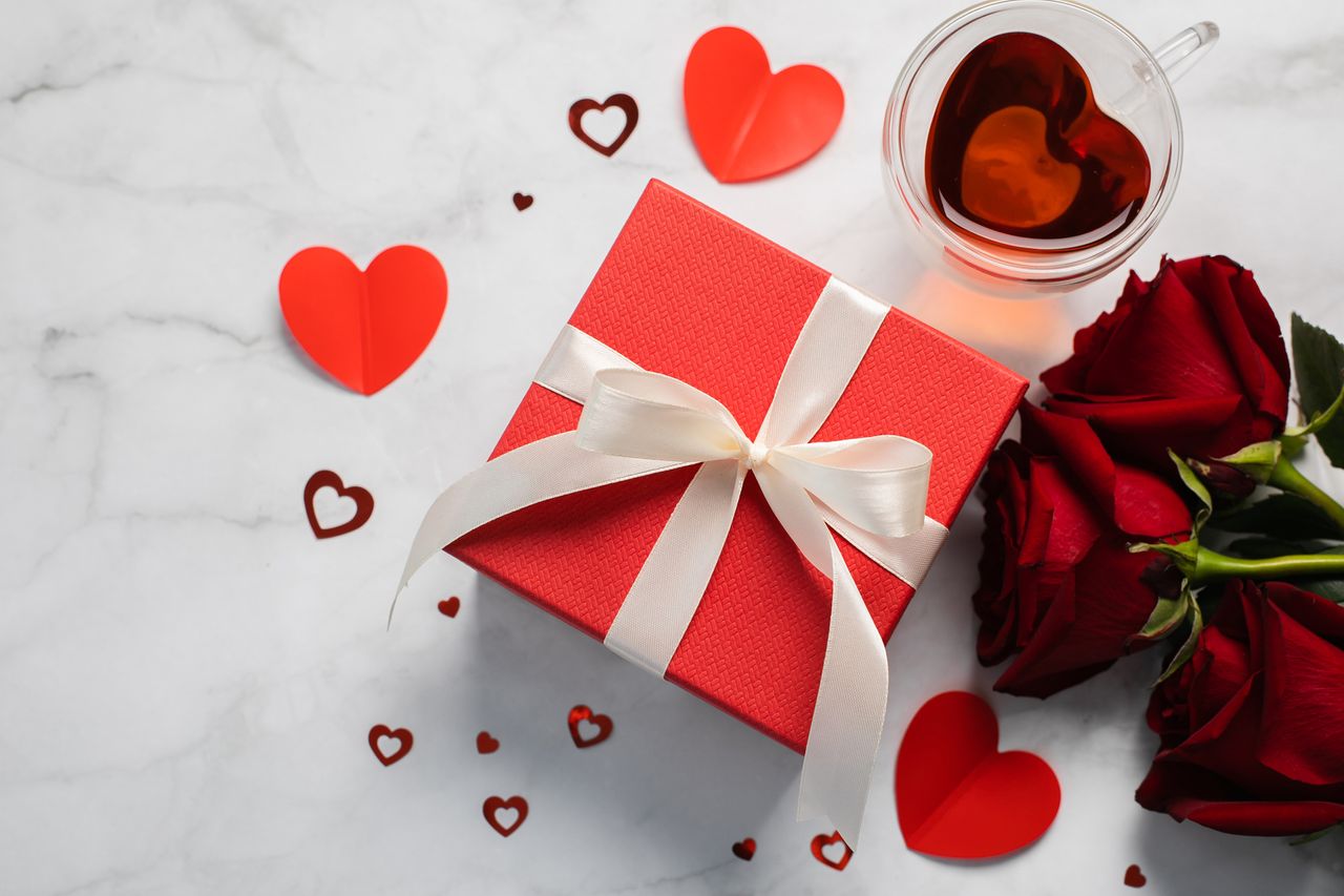 A white marble background with a red present for valentine&#039;s day along with red roses, and red heart confetti.