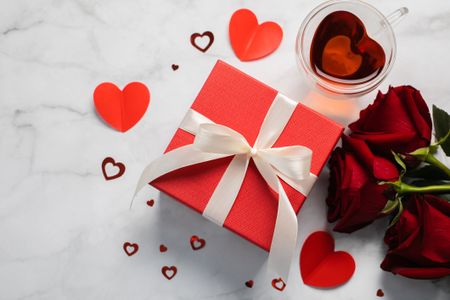 A white marble background with a red present for valentine's day along with red roses, and red heart confetti.