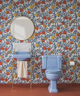 A vibrant bathroom featuring bold, colourful wallpaper adorned with a pomegranate motif in shades of blue, orange, and yellow. The centrepiece is a pale blue pedestal basin with dual brass taps and a towel rail, paired with a matching blue toilet.