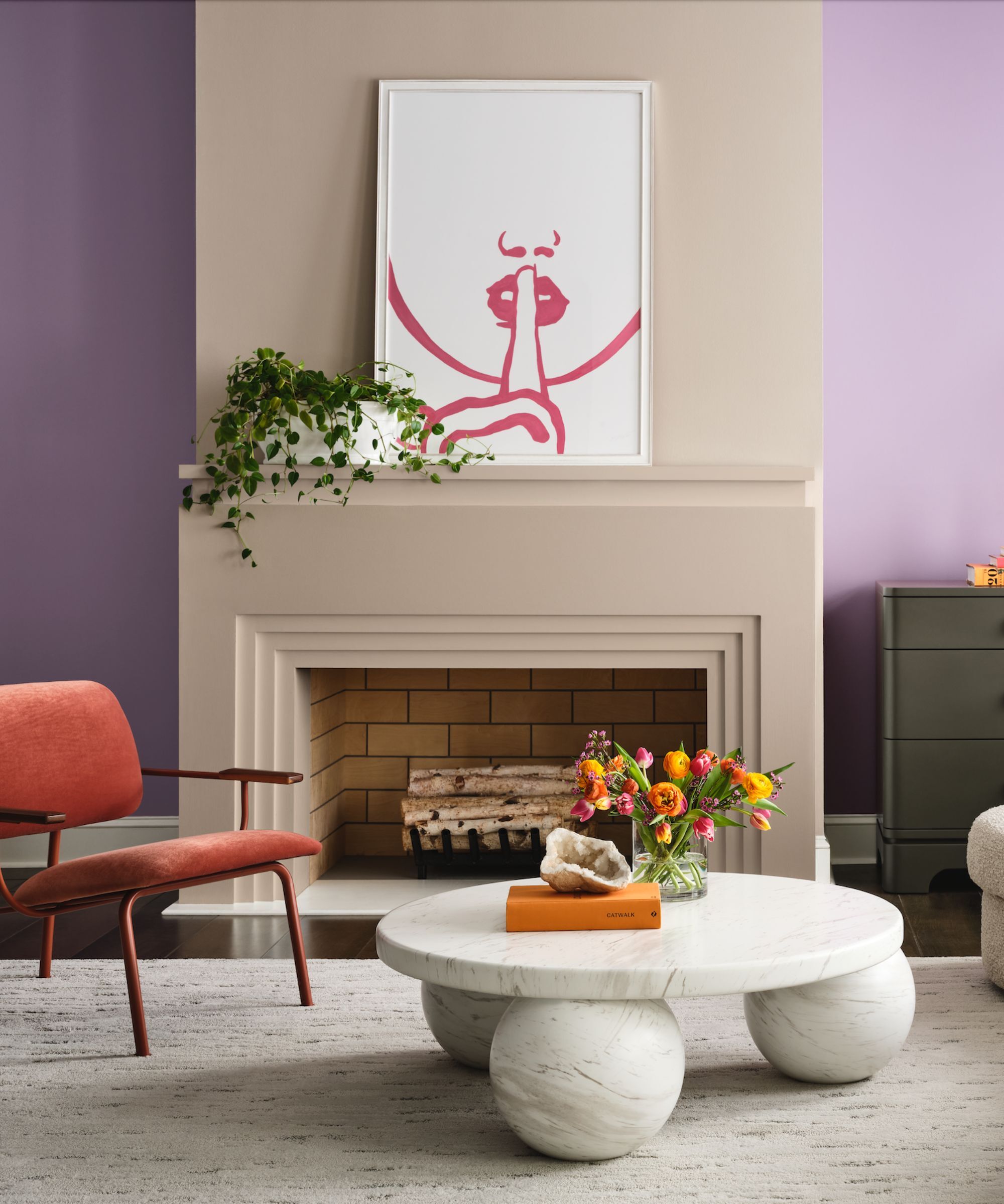A sitting room with purple walls, stone colored fireplace, orange velvet chair and a marble coffee table with spherical feet