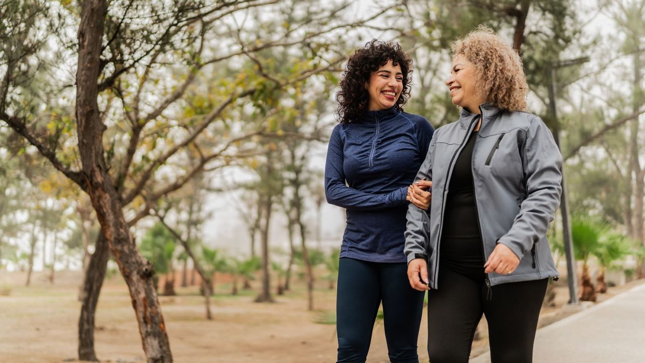 A pair of women in fitness gear walk through a park. One is older, the other is younger, both are smiling as though sharing a joke