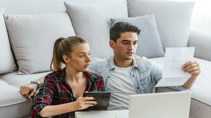 A young couple sit on the floor looking at bills on their computer.