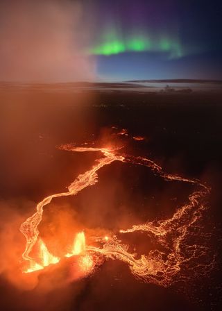 A photograph of green northern lights over an erupting volcano