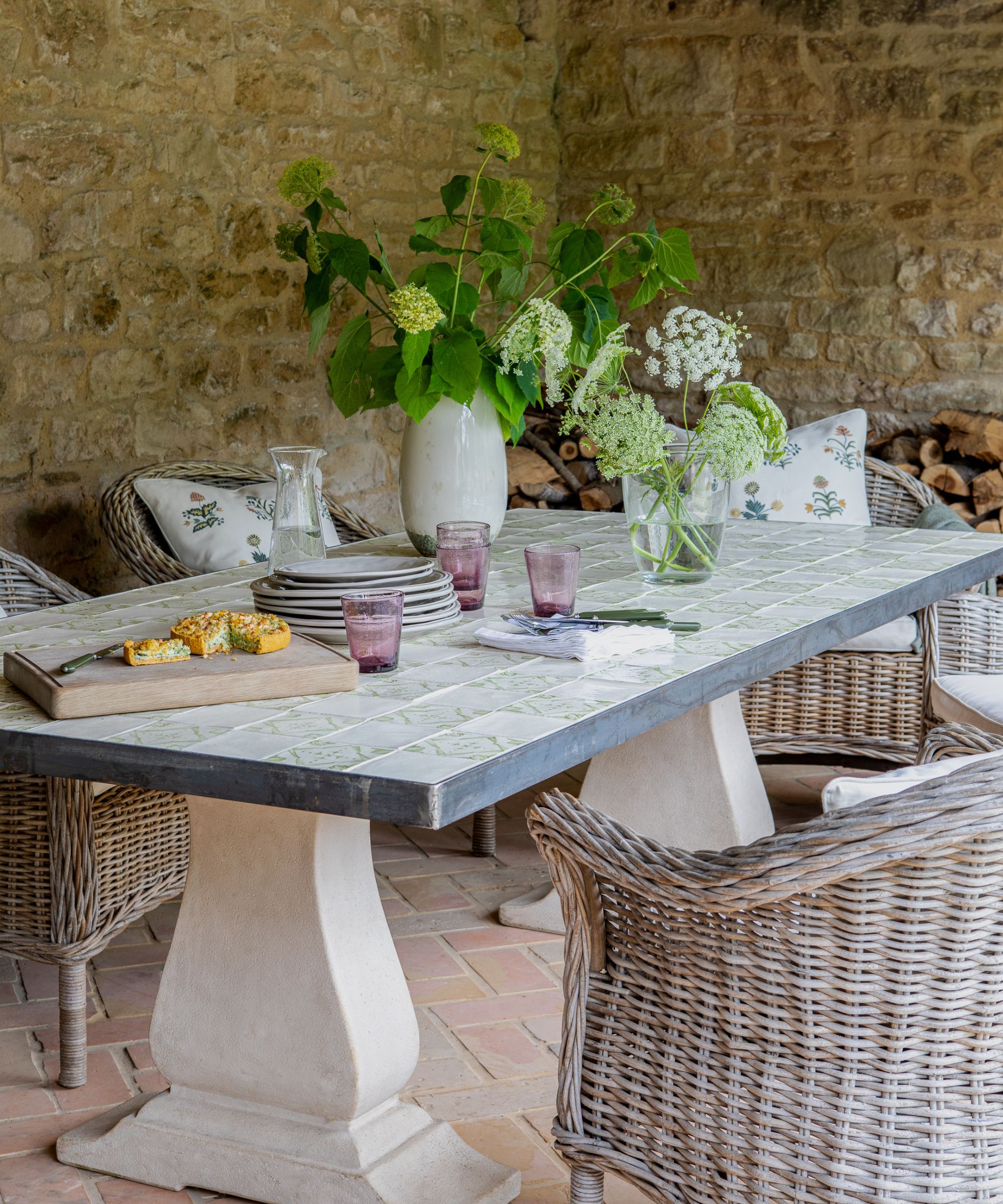 A rustic stone-walled covered outdoor dining area with a tiled-top table on stone pedestals, grey wicker armchairs, purple glasses, wildflower arrangements and terracotta brick flooring.