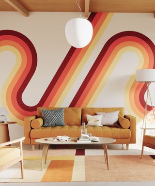 A living room with a white wall with a burgundy and yellow wavy stripe, a light brown couch, a wooden coffee table, and a white rug with orange and brown rectangles on it