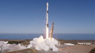 a white and black rocket lifts off from an ocean-side launch pad into a blue sky