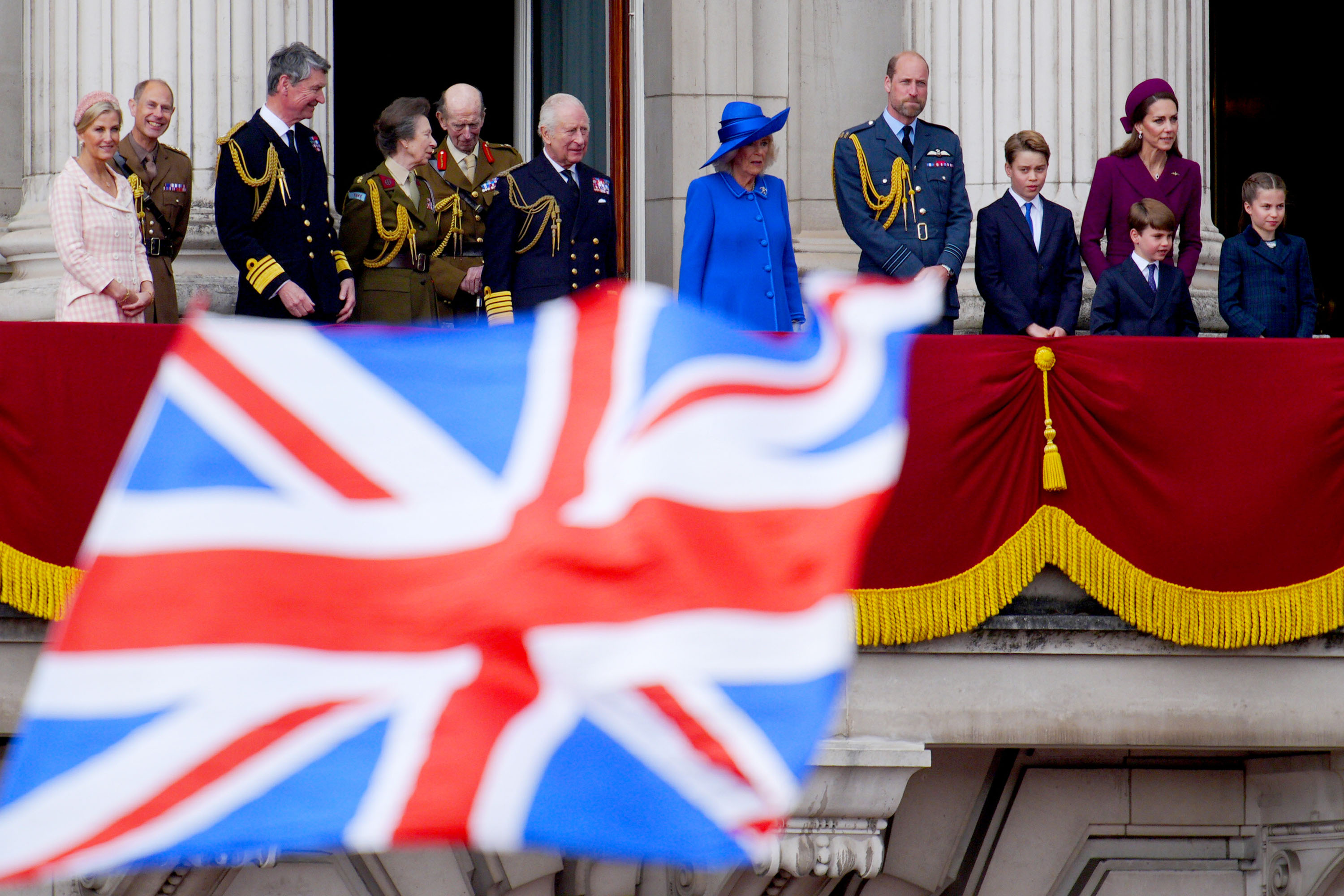 A British flag in front of the balcony at Buckingham Palace, where The King, Queen, Prince William, Princess Kate and other members are standing on VE Day