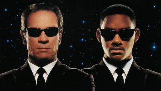 Two men win black suits and a white shirt with a black tie stand side by side while wearing black shades on a starry night background