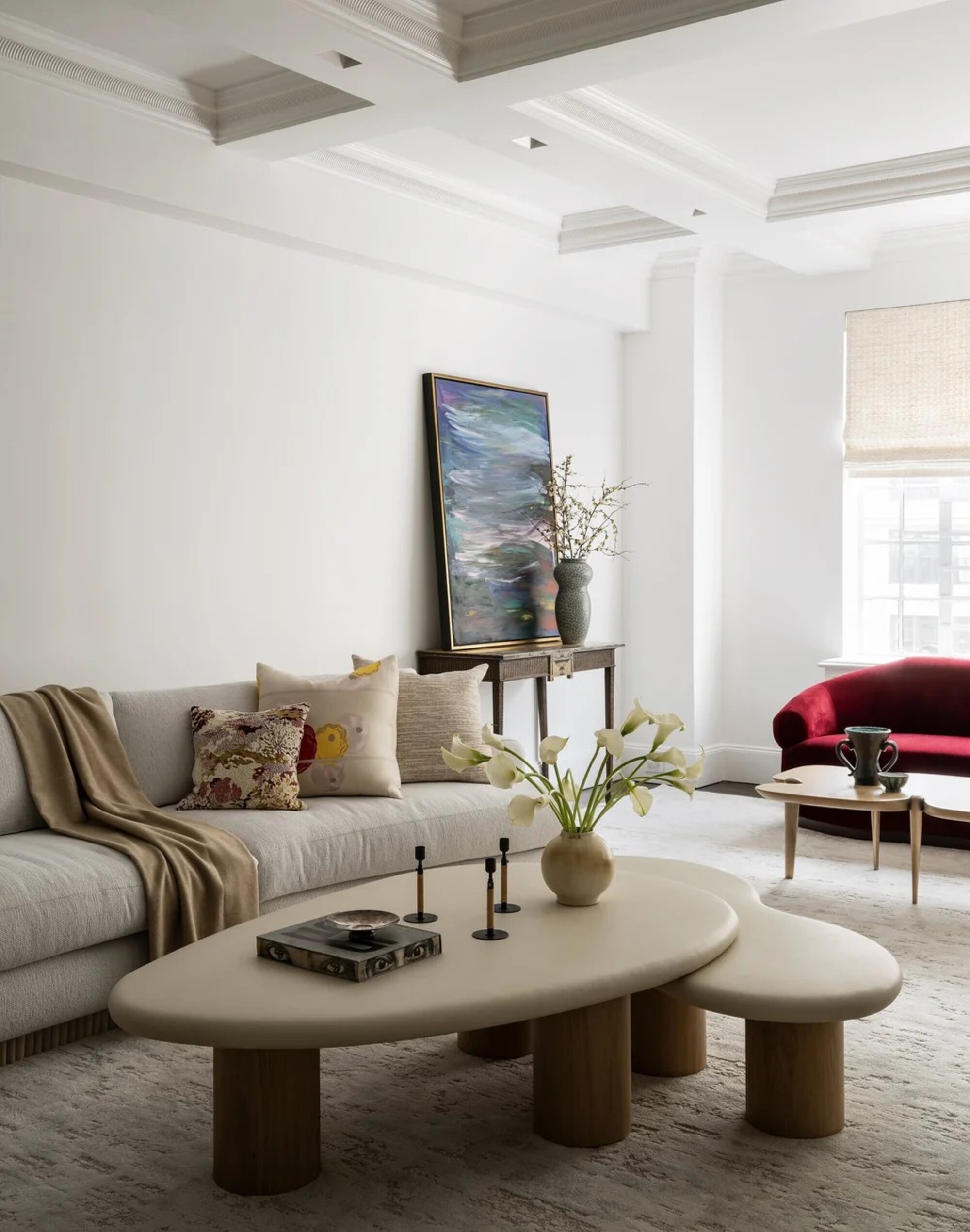 A white, bright living room with a white sofa with cozy throws and a cream nesting coffee table with candlesticks, a vase of lilies, and a trinket tray on it.