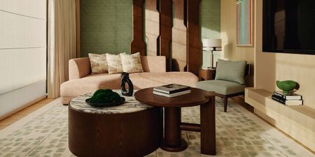 A green and sand-hued hotel suite is filled with contemporary design elements, including two circular wood and marble coffee tables, a voluminous, 1970s-inspired rose couch in sherling, a green and beige sweeping rug, artworks, and print collectibles.