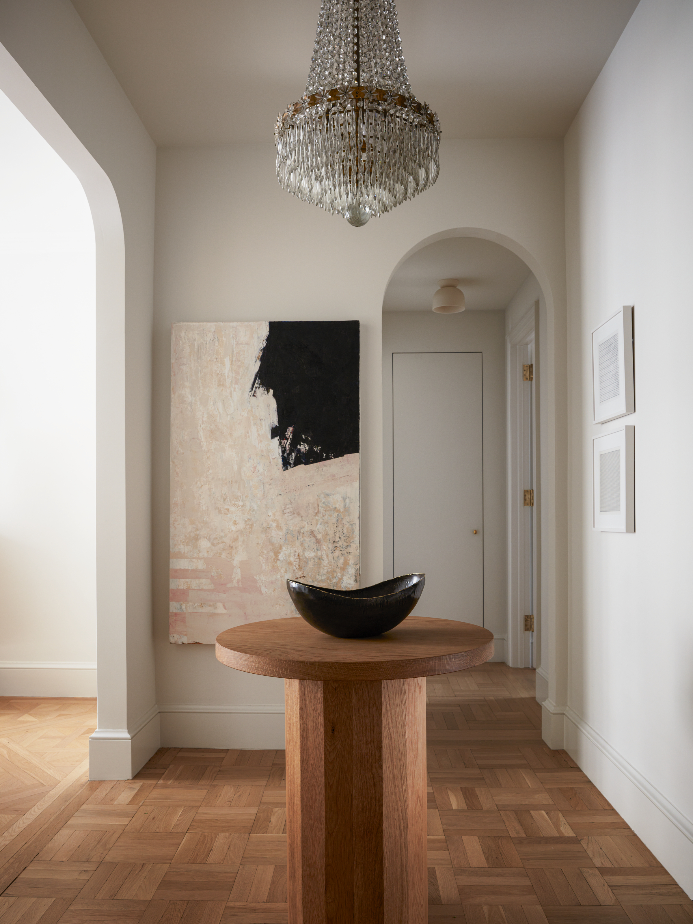Entrance with parquet flooring, small round table in the centre of the room with a black decorative bowl on top, beige and black abstract artwork and a chandelier