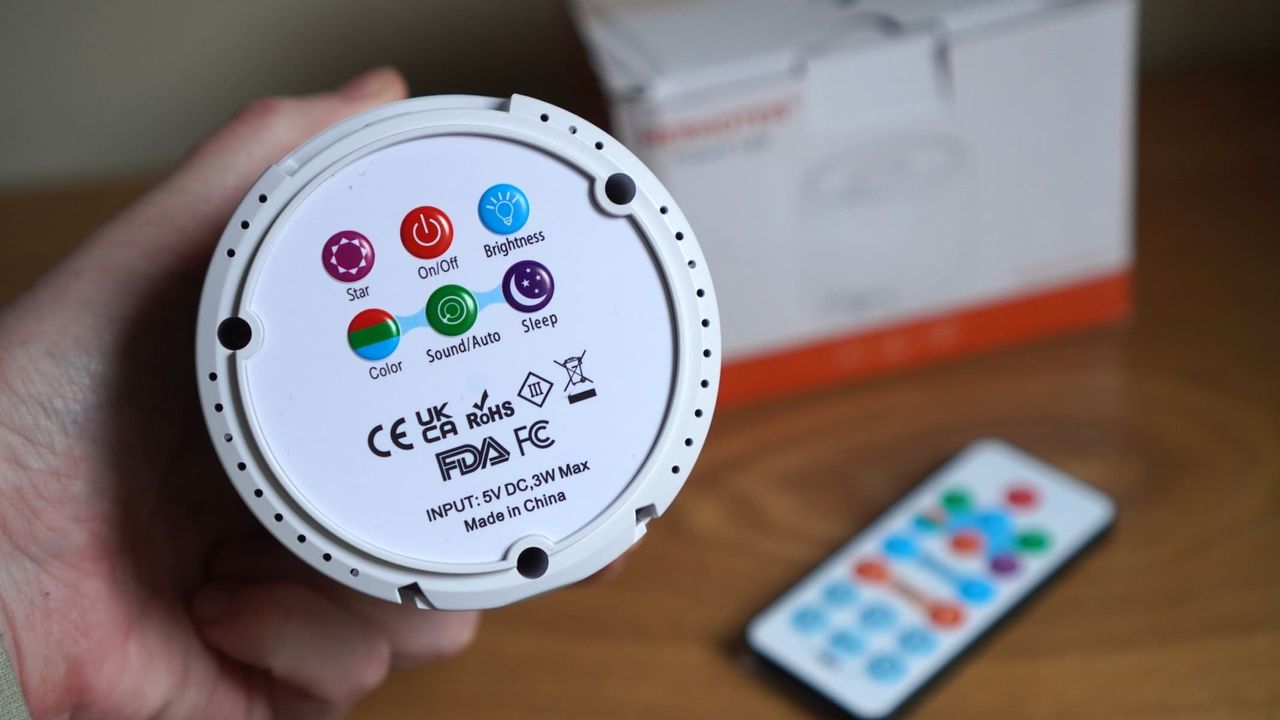 The base of a white spherical projector, being held in a hand, is showing control buttons, with a remote control and a white box in the background