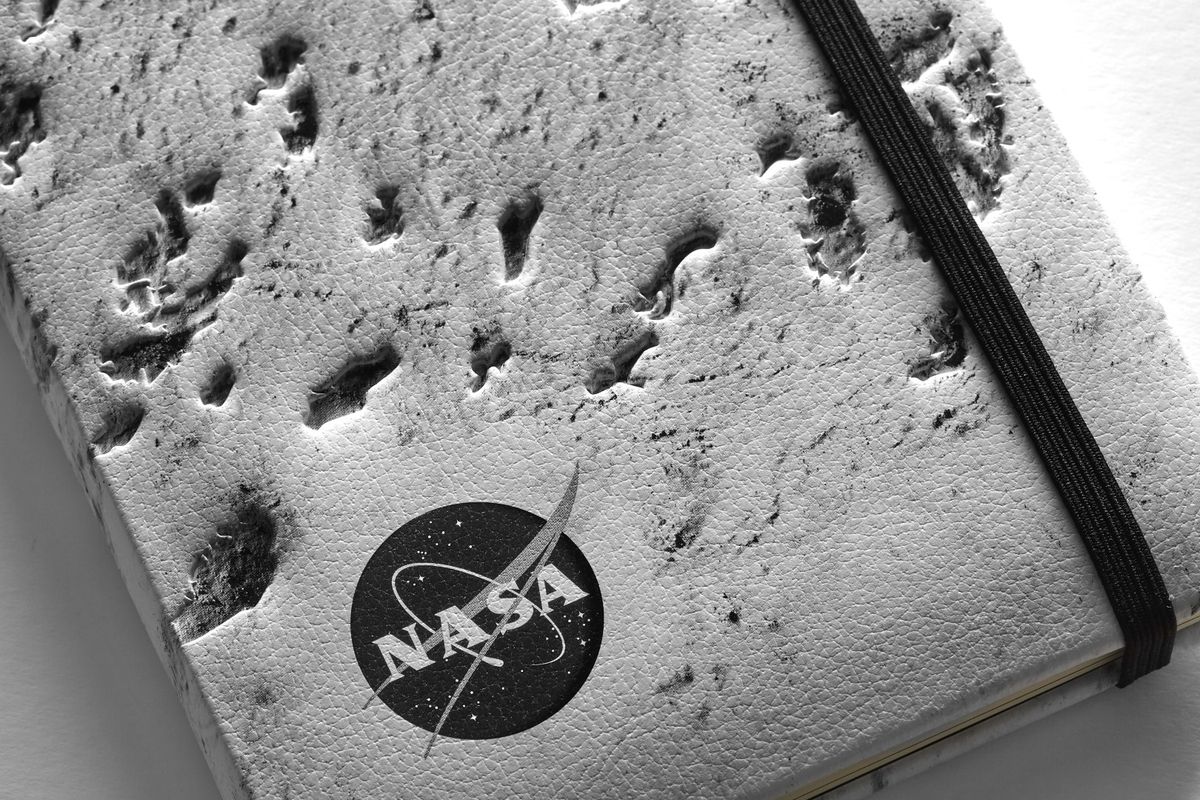 Close up a NASA-inspired Limited Edition Notebook from Moleskine in silver. The cover is a close up of the moon's surface. The notebook has a black elastic closure. A black and white version of the NASA logo is in the bottom left corner.