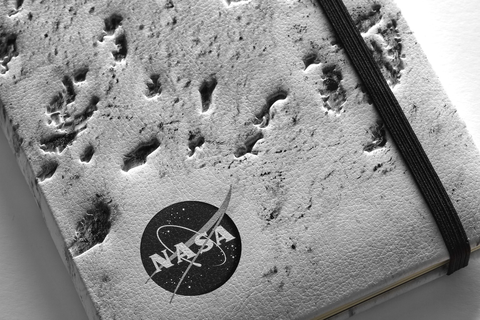 Close up a NASA-inspired Limited Edition Notebook from Moleskine in silver. The cover is a close up of the moon's surface. The notebook has a black elastic closure. A black and white version of the NASA logo is in the bottom left corner.