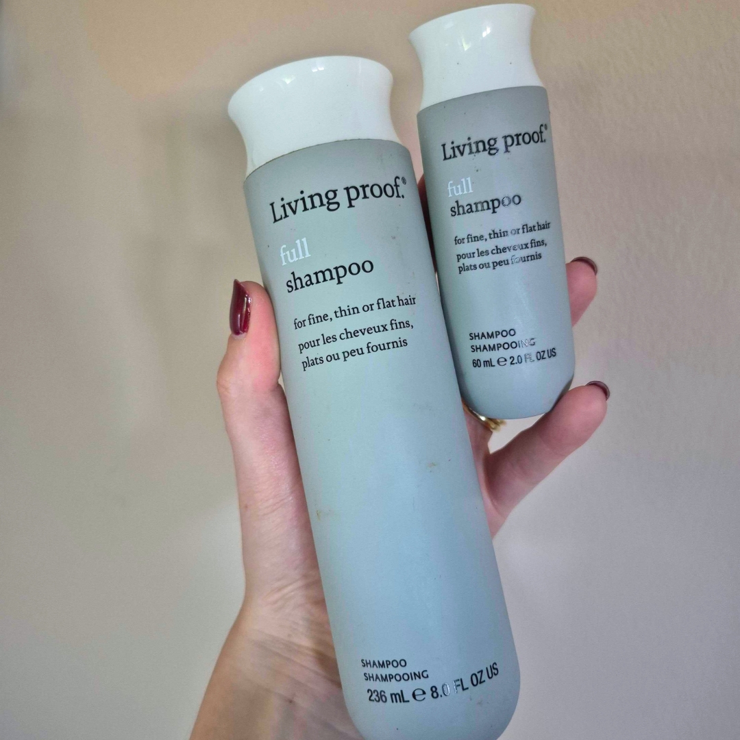 Hand holding two empty bottles of Living Proof Full Shampoo, one normal and travel-sized