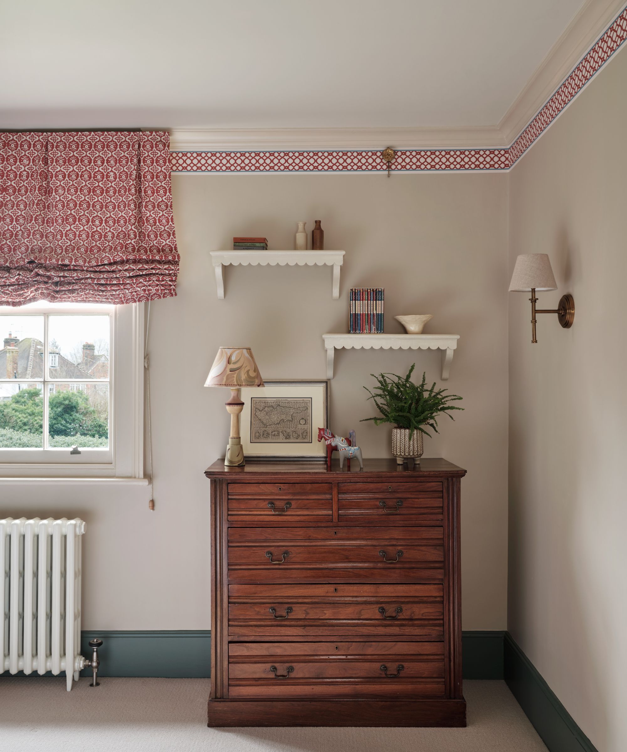 Bedroom with pale grey walls, ceiling trim and matching red blinds, antique wood chest of drawers and sconces