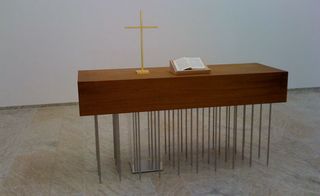 Interior alter space, grey marble effect floor, white wall, wooden block desk with multiple metal rods as leg support, book open and light wood cross on a small stand on the desk top