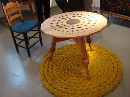 A woven rug underneath a three legged table with an intricate design on the table top. The Roosje chair, a normal wooden chair with a blue cushion seat.