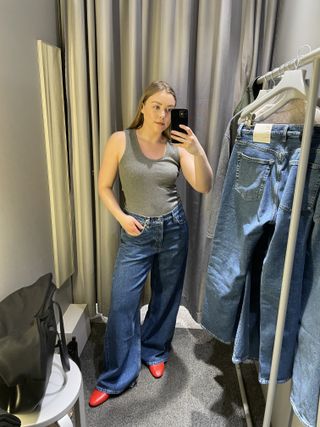 Woman in dressing room wears grey vest, blue jeans and red shoes