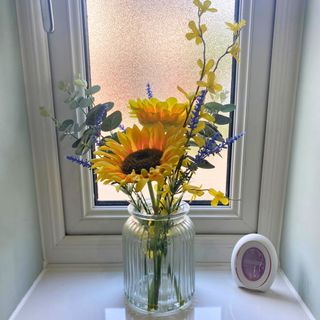 Febreze Small Space air freshener on a small bathroom windowsill next to a ribbed glass plant with sunflowers and spring florals