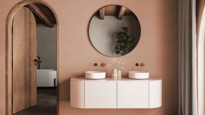 Bathroom vanity showcasing a round mirror on a terracotta pink wall hanging over a white double sink vanity