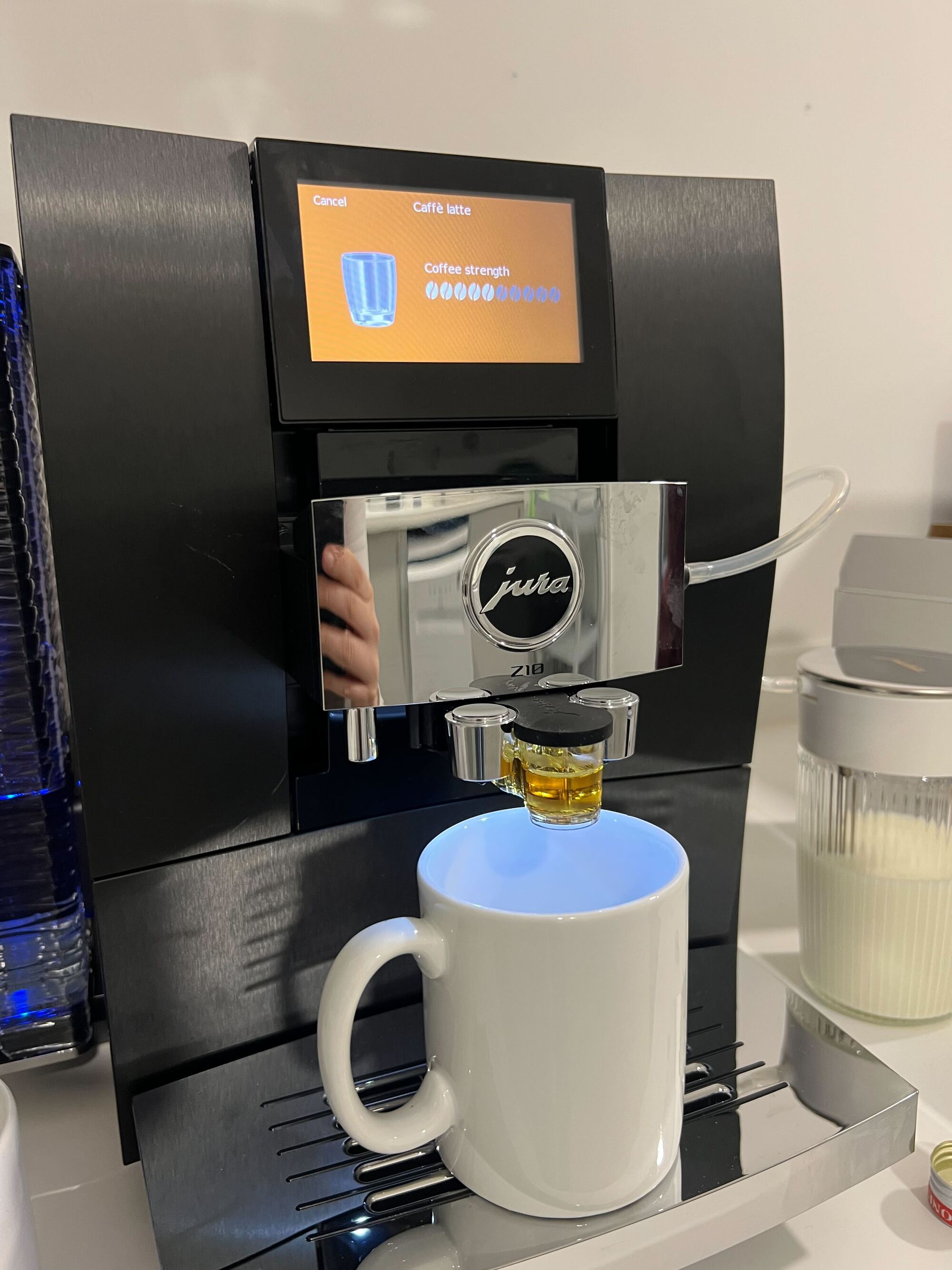 Image of a black and silver jura coffee machine with the syrup function in and a white coffee mug on the drip tray. There is a clear milk container next to the machine.