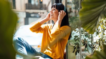 Woman happy and smiling, wearing headphones and sitting near a large window, a few of the ways to boost your energy levels