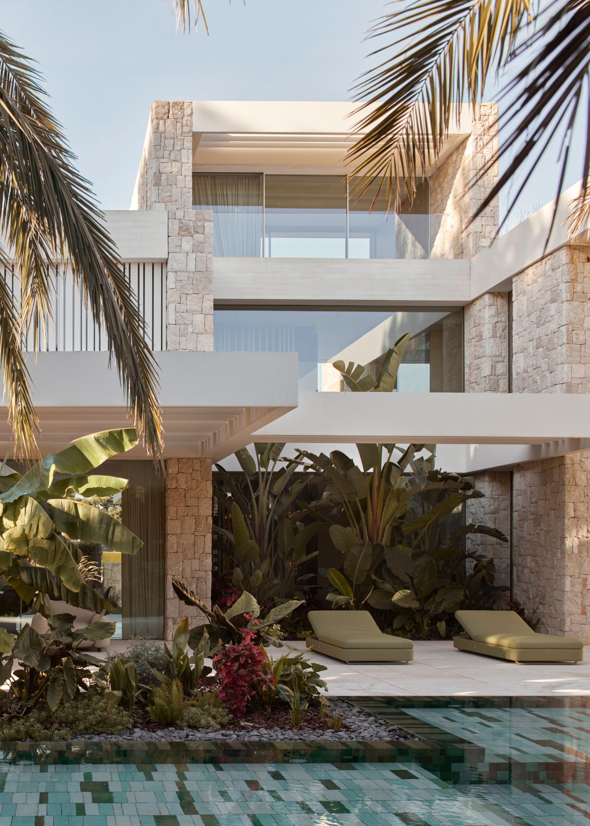 A garden with lush landscaping, a tiled pool, a white stone facade, and green sunbeds