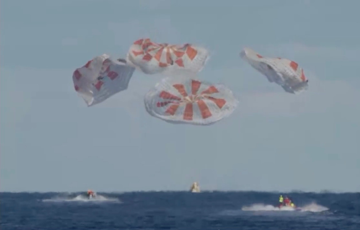 a cone-shaped spacecraft splashing down in the ocean with parachutes deflating above. boats in the water are racing to the spacecraft