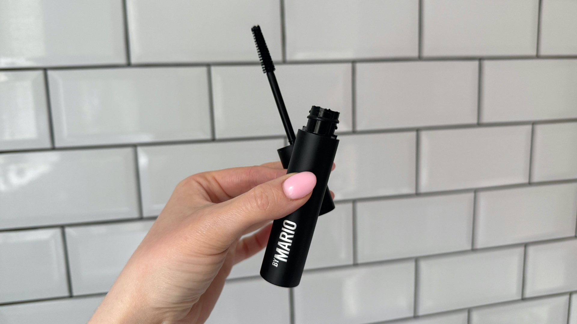 Close up of a handheld view of the Makeup by Mario Master Mascara open with its wand in front of a white tiled background