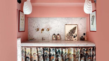 A pink painted bathroom corner. A vanity with a white marble counter and backsplash, gold faucet, and a curtain front. 