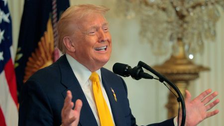 a man in a blue suit with a yellow tie speaks into a microphone in a richly decorated room