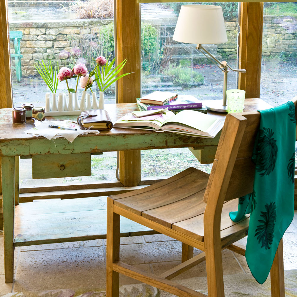 A vintage desk in a wooden conservatory with a wooden chair and green throw