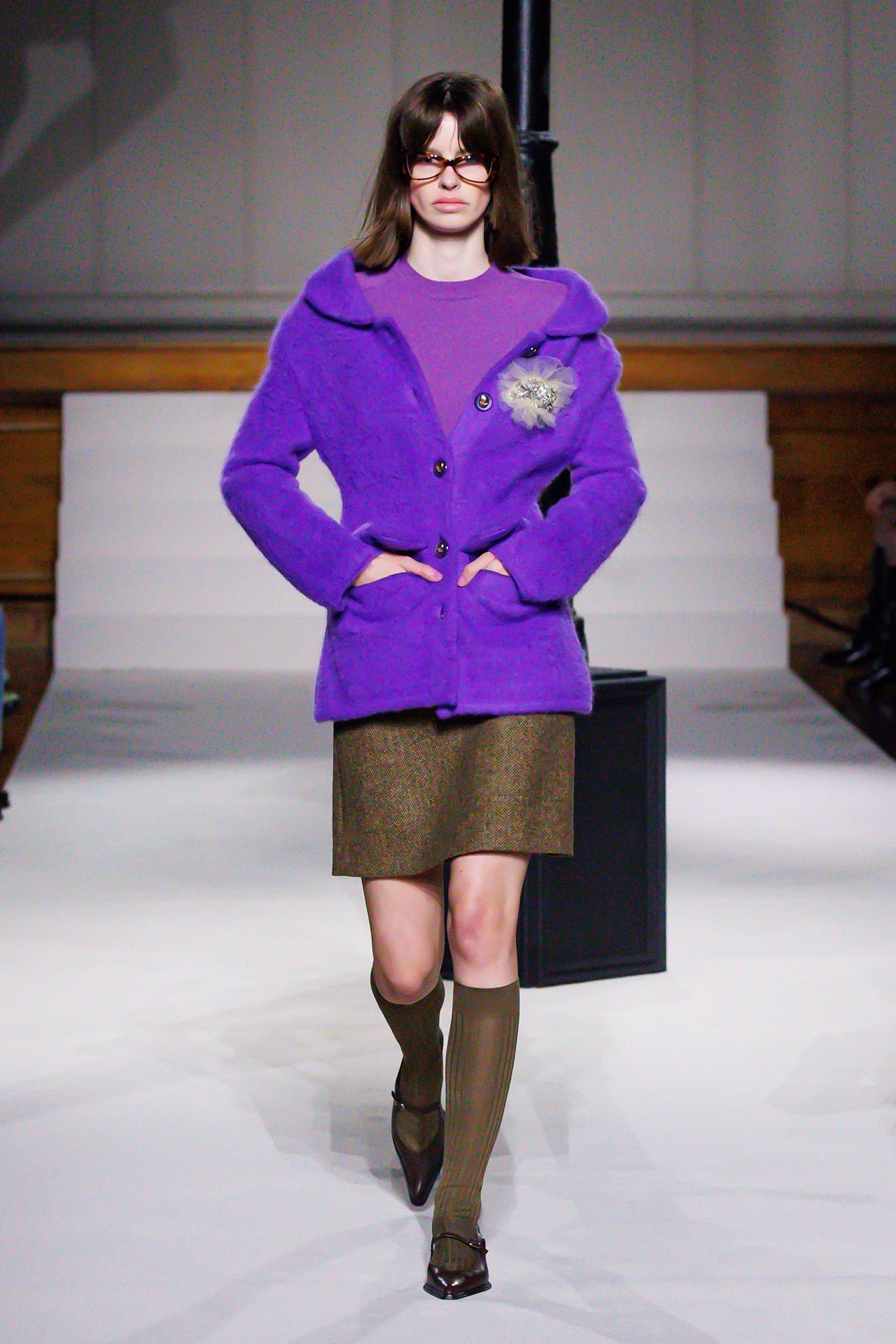 a model on the Kent and Curwen runway wears a purple t-shirt, cardigan and brown skirt and knee high socks with pointed flats.