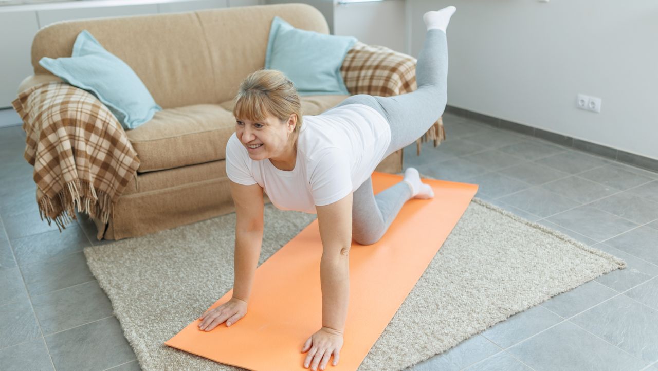 A woman performs a donkey kick at home on a yoga mat. She is on all fours, with her hands on the floor and her arms straight. One knee rests bent on the floor while her other leg is in the air. The elevated leg is bent at the knee, so the thigh is parallel to the floor and the foot pointing towards the ceiling. Behind her is a couch and a white washed wall.