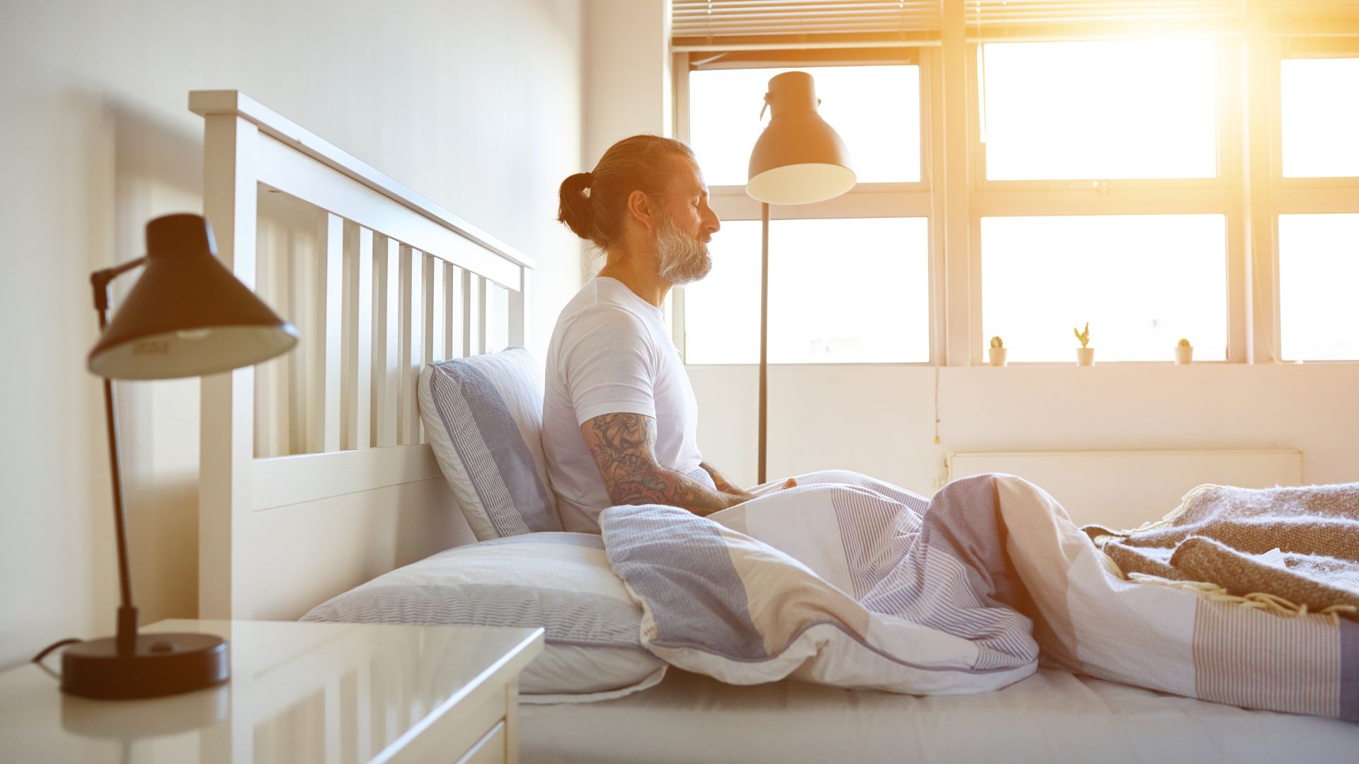 A man sits in bed with his eyes closed and legs crossed in front of him. He is practising a form of meditation.