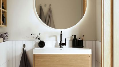 ikea bathroom with timber-look vanity, round mirror with LED light, white tiles, timber trims, timber shelving, black hand soap pump, and small vase with flowers