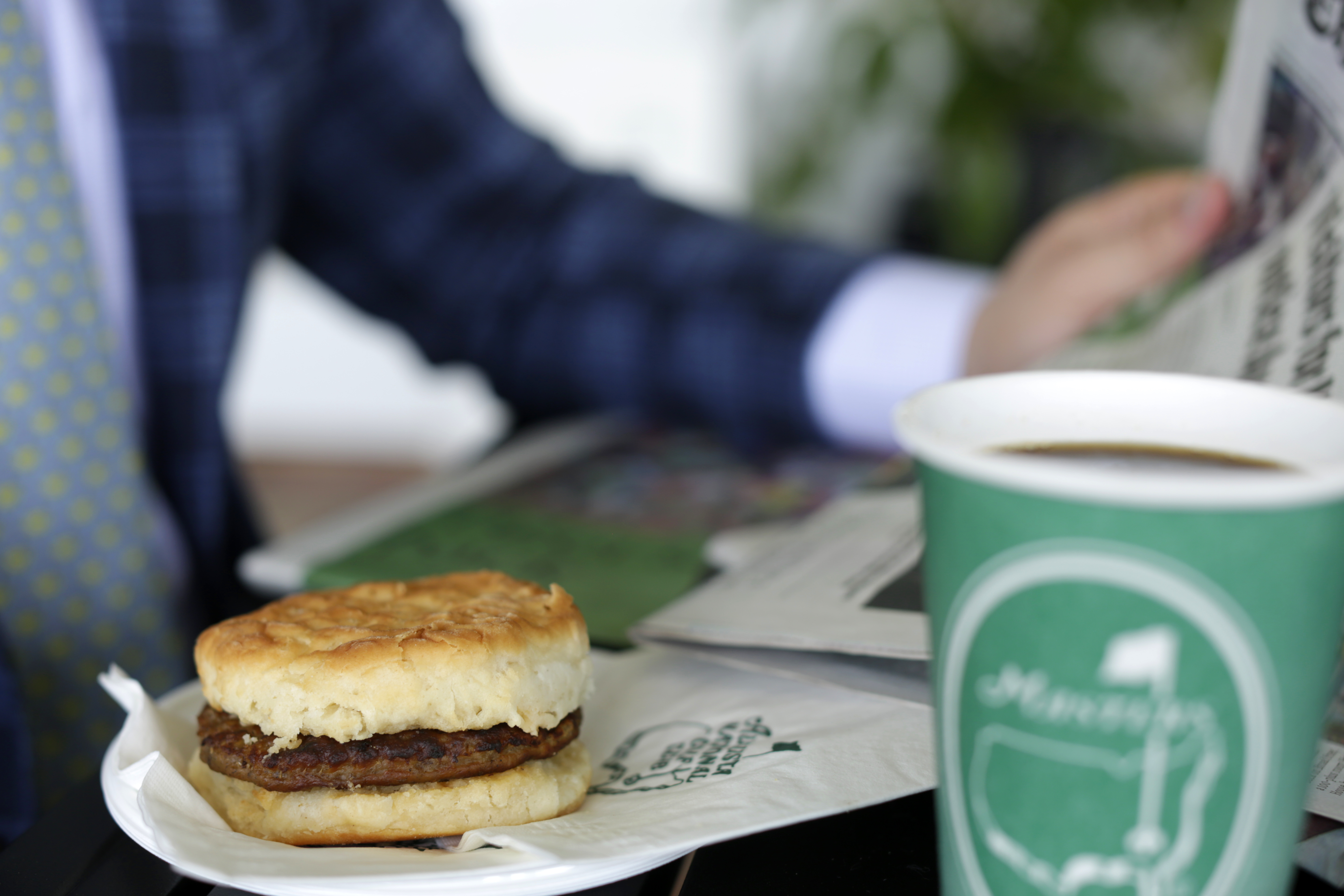 A sausage biscuit and green Masters coffee cup on a table while someone reads the newspaper