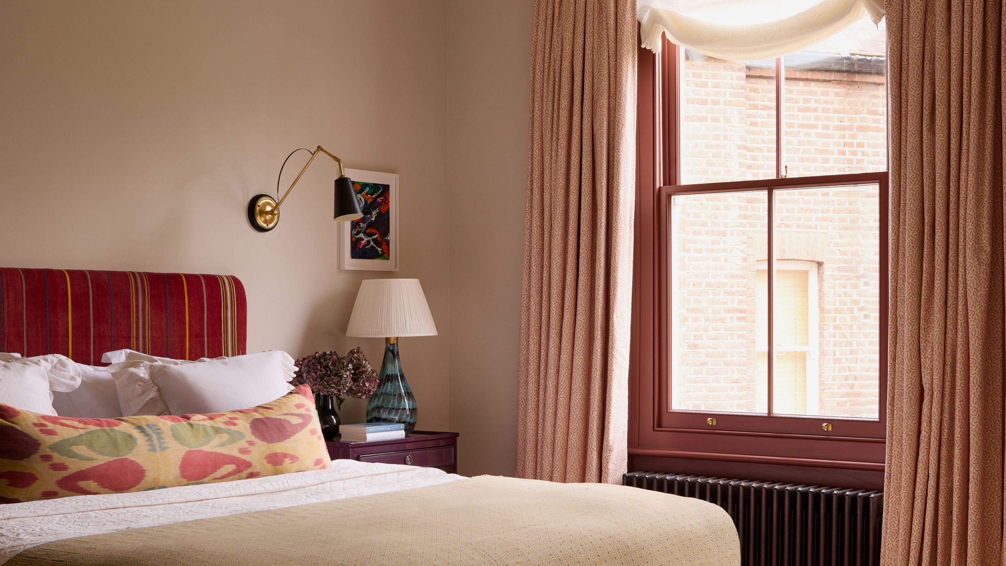 Image of a bedroom with beige walls and a dark red window frame with pink curtains. The bed is red with white and beige bedding.