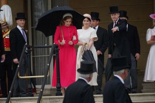 Princess Beatrice wearing a red dress and holding a black umbrella standing at the top of a staircase next to Princess Eugenie in a white short sleeve dress