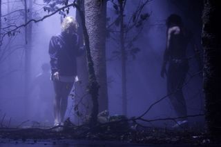 A man and a woman are standing in a mist-covered forest that has a purple haze.