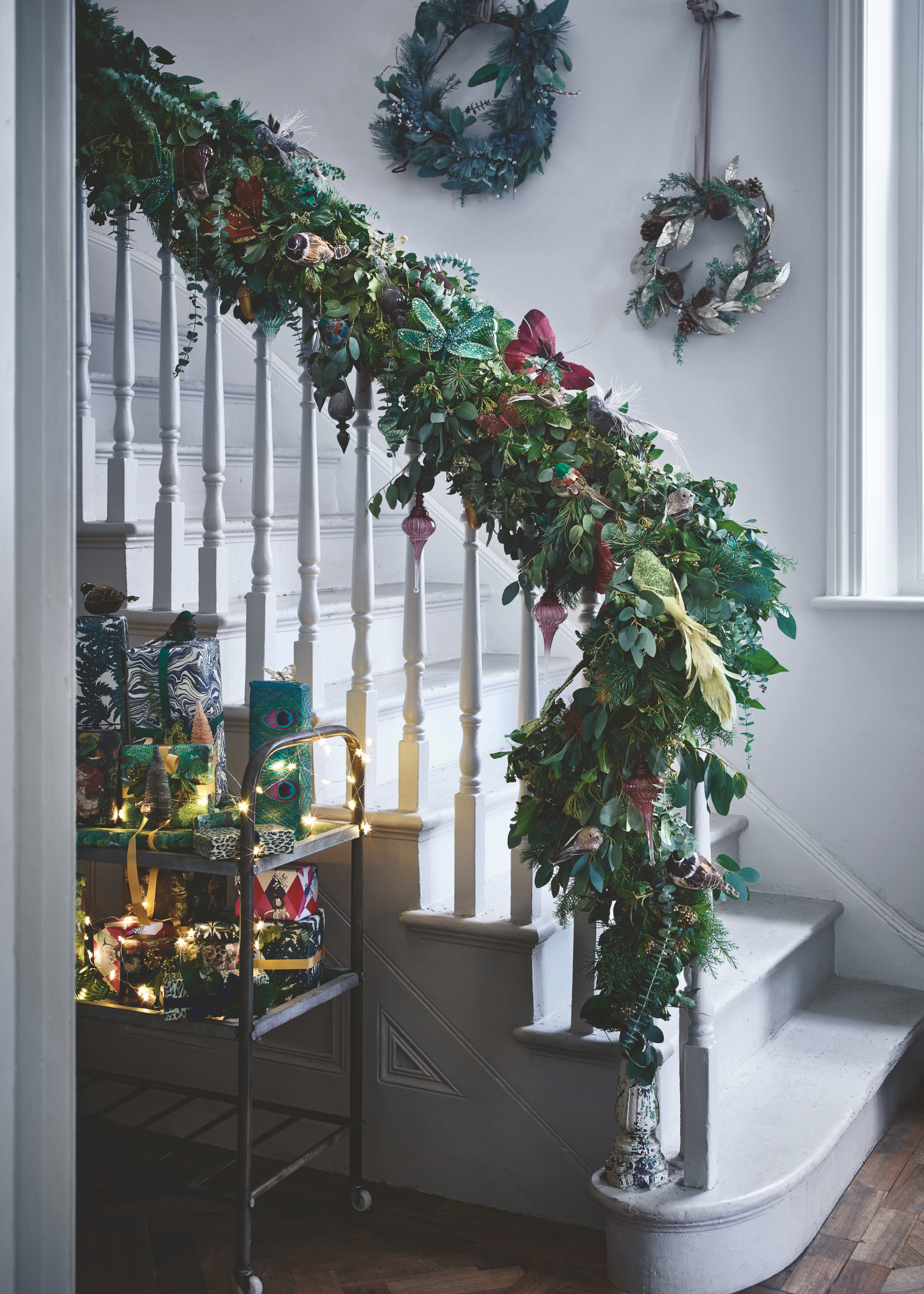 An entryway is covered in decorations from the cabinet at the bottom of the stairs to the ornate garland on the stair rail and the two wreaths hanging from the wall along the side of the stairs