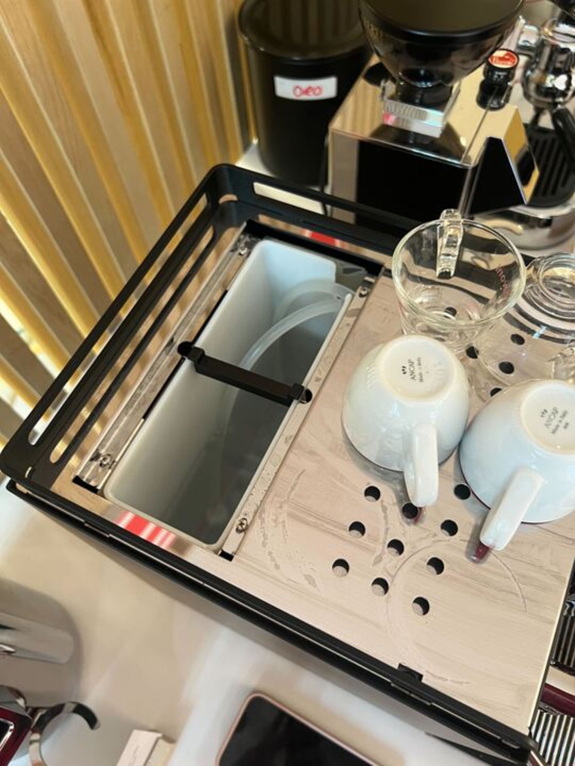 The top of a silver coffee machine that has coffee mugs on it and the water tank lid open.